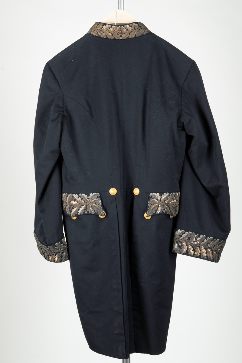 Paraduniformsrock till landshövdingeuniform. Mörkblå frackrock, v-ringad med ståkrage. Kragen, ärmslag och ficklock har broderade eklöv i gulmetalltråd. Knäppt fram med en hyska och hake. Åtta stycken garneringsknappar med motsvarande markerade knapphål. Knappar i gylmetall med krönt örn i hertigkrona. Livsömmar och skörtveck bak. Två knappar samt två ficklock med vardera tre knappar i midjesömmen bak. Tvåsömsärm med blindslag. Vadderad, foder av svart atlassiden. Fickor i skörtets foder.