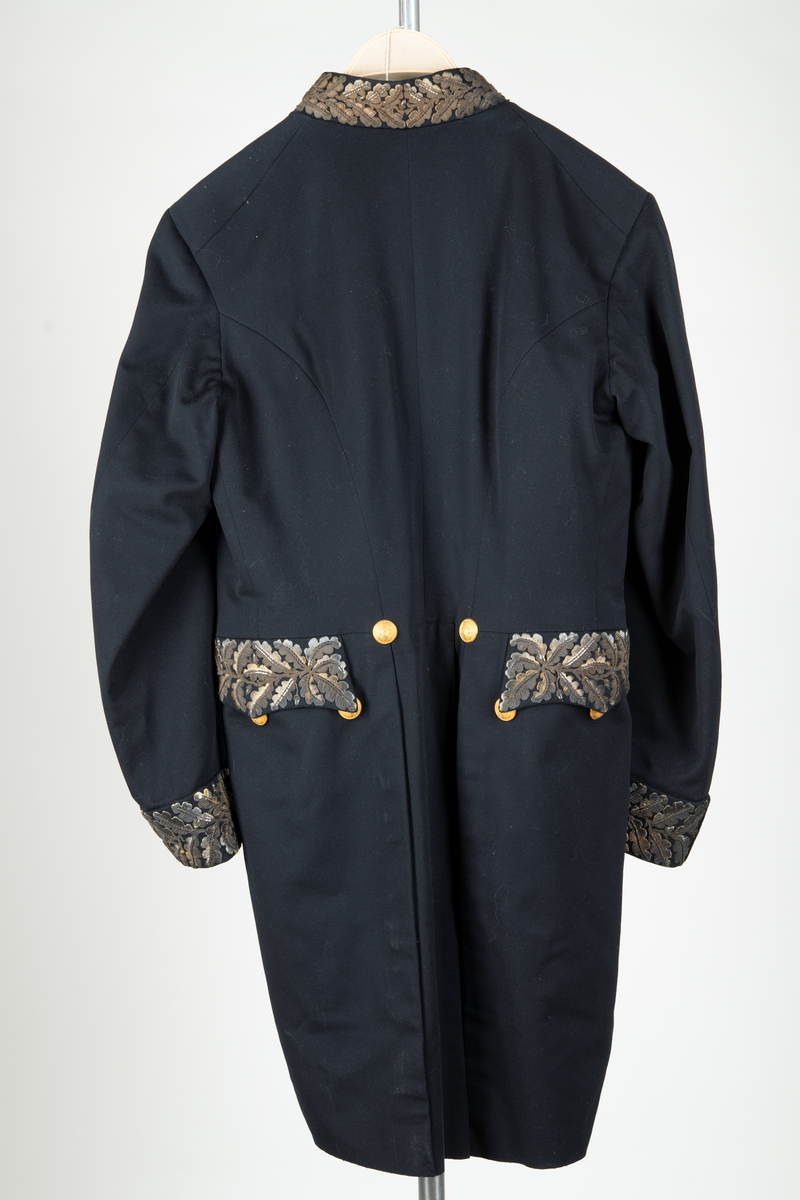 Paraduniformsrock till landshövdingeuniform. Mörkblå frackrock, v-ringad med ståkrage. Kragen, ärmslag och ficklock har broderade eklöv i gulmetalltråd. Knäppt fram med en hyska och hake. Åtta stycken garneringsknappar med motsvarande markerade knapphål. Knappar i gylmetall med krönt örn i hertigkrona. Livsömmar och skörtveck bak. Två knappar samt två ficklock med vardera tre knappar i midjesömmen bak. Tvåsömsärm med blindslag. Vadderad, foder av svart atlassiden. Fickor i skörtets foder.