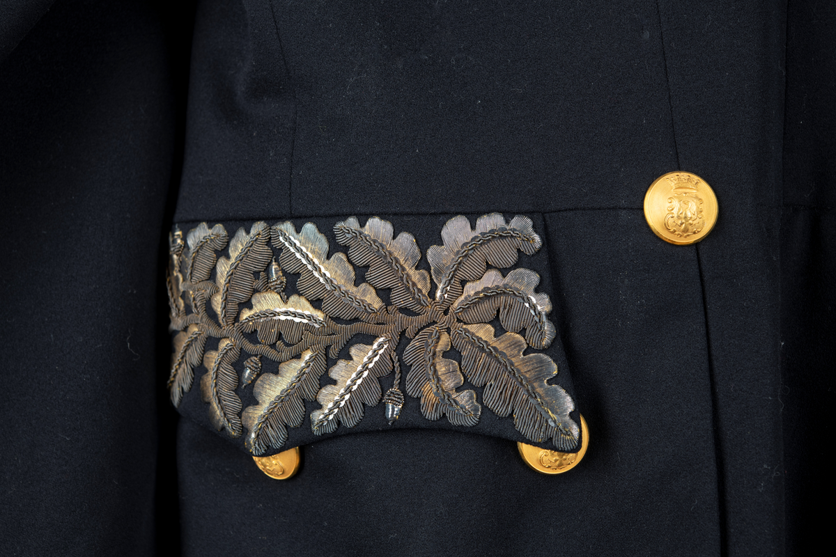 Paraduniformsrock till landshövdingeuniform. Mörkblå frackrock, v-ringad med ståkrage. Kragen, ärmslag och ficklock har broderade eklöv i gulmetalltråd. Knäppt fram med en hyska och hake. Åtta stycken garneringsknappar med motsvarande markerade knapphål. Knappar i gylmetall med krönt örn i hertigkrona. Livsömmar och skörtveck bak. Två knappar samt två ficklock med vardera tre knappar i midjesömmen bak. Tvåsömsärm med blindslag. Vadderad, foder av svart atlassiden. Fickor i skörtets foder.