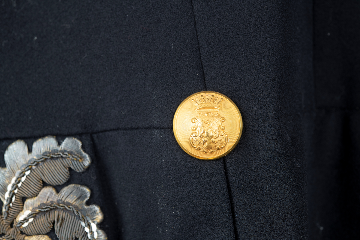 Paraduniformsrock till landshövdingeuniform. Mörkblå frackrock, v-ringad med ståkrage. Kragen, ärmslag och ficklock har broderade eklöv i gulmetalltråd. Knäppt fram med en hyska och hake. Åtta stycken garneringsknappar med motsvarande markerade knapphål. Knappar i gylmetall med krönt örn i hertigkrona. Livsömmar och skörtveck bak. Två knappar samt två ficklock med vardera tre knappar i midjesömmen bak. Tvåsömsärm med blindslag. Vadderad, foder av svart atlassiden. Fickor i skörtets foder.