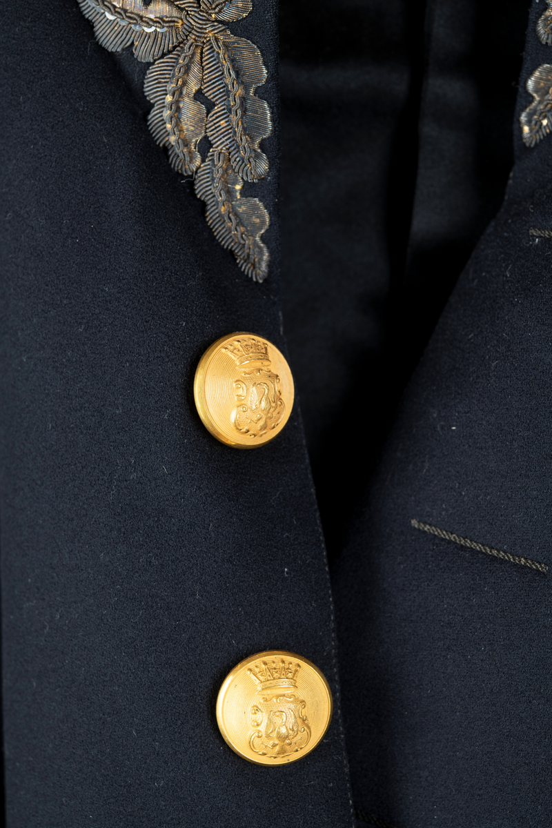 Paraduniformsrock till landshövdingeuniform. Mörkblå frackrock, v-ringad med ståkrage. Kragen, ärmslag och ficklock har broderade eklöv i gulmetalltråd. Knäppt fram med en hyska och hake. Åtta stycken garneringsknappar med motsvarande markerade knapphål. Knappar i gylmetall med krönt örn i hertigkrona. Livsömmar och skörtveck bak. Två knappar samt två ficklock med vardera tre knappar i midjesömmen bak. Tvåsömsärm med blindslag. Vadderad, foder av svart atlassiden. Fickor i skörtets foder.