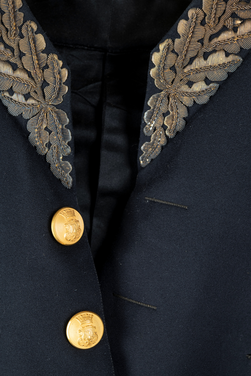 Paraduniformsrock till landshövdingeuniform. Mörkblå frackrock, v-ringad med ståkrage. Kragen, ärmslag och ficklock har broderade eklöv i gulmetalltråd. Knäppt fram med en hyska och hake. Åtta stycken garneringsknappar med motsvarande markerade knapphål. Knappar i gylmetall med krönt örn i hertigkrona. Livsömmar och skörtveck bak. Två knappar samt två ficklock med vardera tre knappar i midjesömmen bak. Tvåsömsärm med blindslag. Vadderad, foder av svart atlassiden. Fickor i skörtets foder.