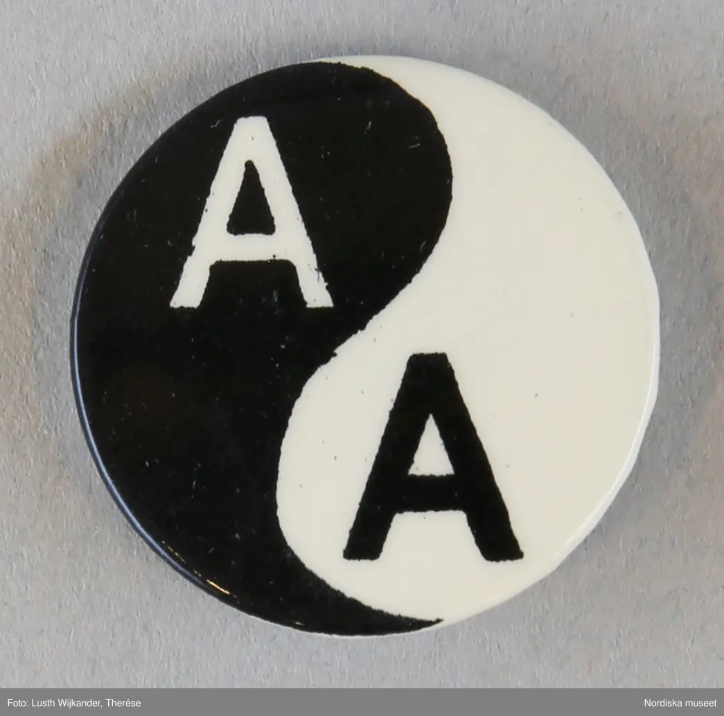 Huvudliggaren:
"Rockslagsmärke, metall. Runt svart/vitt, 2 st, yin/yang märke med monogram AA (anti apartheid). Gåva av sydafrikan till givaren 1961 under vistelse i England. Nål, krylle. Använd i Stockholm.
Margit Forsberg f. 1921 bodde under en kortare tid sommaren 1961 i England i samma familj som en sydafrikan. Han berättade om förhållandena för de svarta i sitt hemland, vilket grep henne djupt. Hon fick 2 märken av honom, som sedan bars hemma i Stockholm.
/Berit Eldvik 2009-12-18