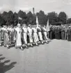 Jubileumsutställningen. Invigningen. 
Ett antal kvinnor i s.k. husmorsdräkt (tillhörande Husmodersförbundet) tågar in på planen framför hedersläktaren.