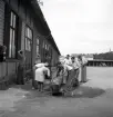 Midsommardagen 1942 vid kreatursstallarna. Ett antal yngre personer har samlats för morgontvättning vid en vattenho utanför en lång träbyggnad. 
I bakgrunden skymtar en bil och tornet på dåv. Dövstumskolan.