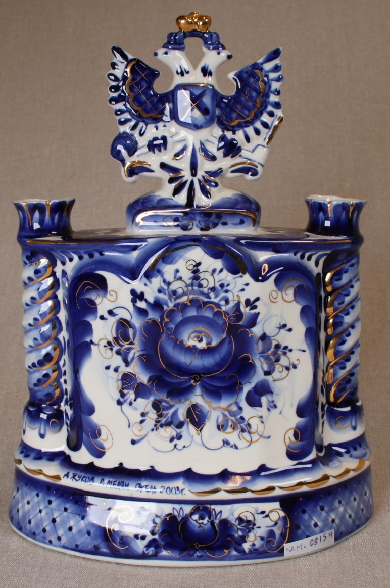 porselensur, hvitt med dekor i blått og gull. IPAs logo i front.
Dobbelørn med gullkrone på toppen.