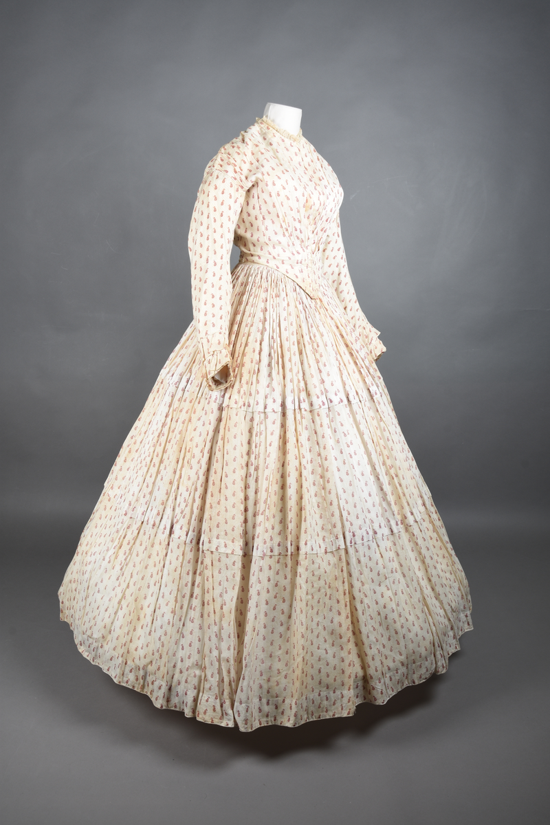 Bomullskjole i to deler, laget av voile med hvit bunn og små strøbuketter i burgunderfarge, hvor prikkene delvis er falmet bort. Skjørtet er sterkt rynket og festet til en smal linning av grov lin, med to legg nederst og en kant med smal bomullsnor. Livet har lange ermer, rund halsringning uten krage, og en skjult hekteåpning som går helt ned. Foran og bak går livet ned i en spiss, med rynkefolder fra skuldrene ned mot spissene. Livet er helfôret, og fôret går inn i ermene. Halsen er kantet med en liten kniplingsrysj, og det er to stoffknapper ved hvert håndledd. Hemper for oppheng er innsydd i skuldersømmene, og det er opphengshemper sydd inn i skjørtets linning. Kjolen er håndsydd.