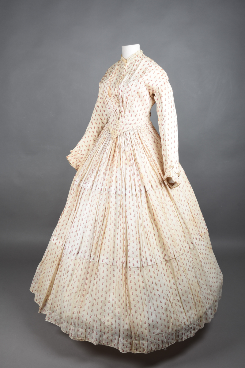 Bomullskjole i to deler, laget av voile med hvit bunn og små strøbuketter i burgunderfarge, hvor prikkene delvis er falmet bort. Skjørtet er sterkt rynket og festet til en smal linning av grov lin, med to legg nederst og en kant med smal bomullsnor. Livet har lange ermer, rund halsringning uten krage, og en skjult hekteåpning som går helt ned. Foran og bak går livet ned i en spiss, med rynkefolder fra skuldrene ned mot spissene. Livet er helfôret, og fôret går inn i ermene. Halsen er kantet med en liten kniplingsrysj, og det er to stoffknapper ved hvert håndledd. Hemper for oppheng er innsydd i skuldersømmene, og det er opphengshemper sydd inn i skjørtets linning. Kjolen er håndsydd.