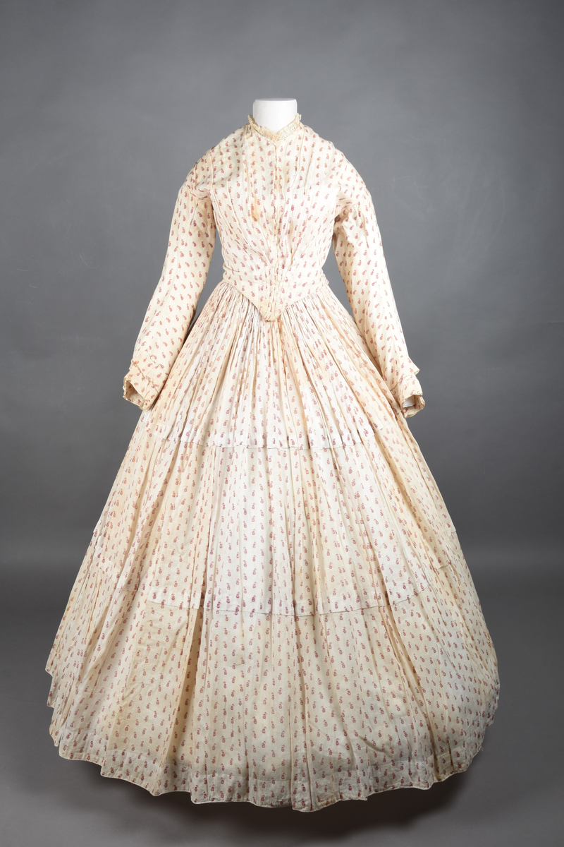 Bomullskjole i to deler, laget av voile med hvit bunn og små strøbuketter i burgunderfarge, hvor prikkene delvis er falmet bort. Skjørtet er sterkt rynket og festet til en smal linning av grov lin, med to legg nederst og en kant med smal bomullsnor. Livet har lange ermer, rund halsringning uten krage, og en skjult hekteåpning som går helt ned. Foran og bak går livet ned i en spiss, med rynkefolder fra skuldrene ned mot spissene. Livet er helfôret, og fôret går inn i ermene. Halsen er kantet med en liten kniplingsrysj, og det er to stoffknapper ved hvert håndledd. Hemper for oppheng er innsydd i skuldersømmene, og det er opphengshemper sydd inn i skjørtets linning. Kjolen er håndsydd.