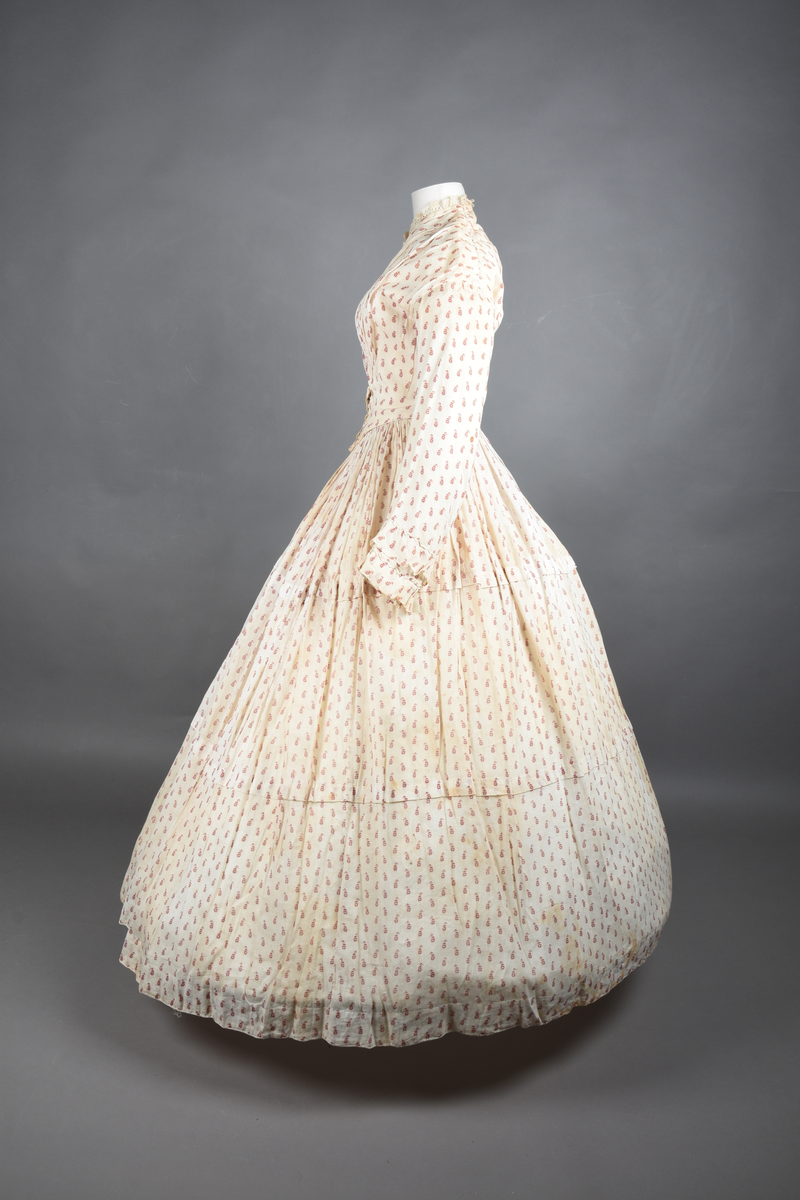 Bomullskjole i to deler, laget av voile med hvit bunn og små strøbuketter i burgunderfarge, hvor prikkene delvis er falmet bort. Skjørtet er sterkt rynket og festet til en smal linning av grov lin, med to legg nederst og en kant med smal bomullsnor. Livet har lange ermer, rund halsringning uten krage, og en skjult hekteåpning som går helt ned. Foran og bak går livet ned i en spiss, med rynkefolder fra skuldrene ned mot spissene. Livet er helfôret, og fôret går inn i ermene. Halsen er kantet med en liten kniplingsrysj, og det er to stoffknapper ved hvert håndledd. Hemper for oppheng er innsydd i skuldersømmene, og det er opphengshemper sydd inn i skjørtets linning. Kjolen er håndsydd.