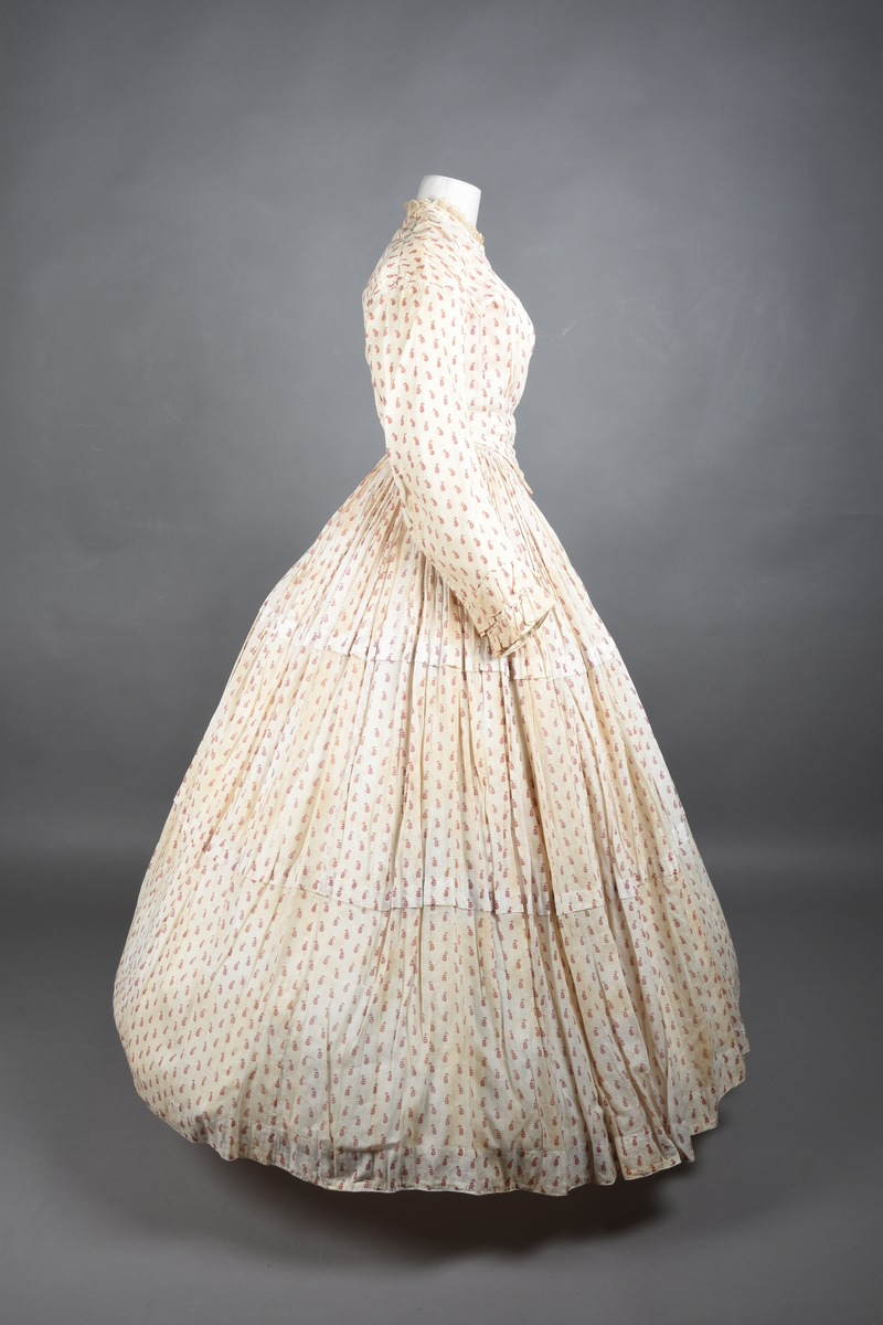 Bomullskjole i to deler, laget av voile med hvit bunn og små strøbuketter i burgunderfarge, hvor prikkene delvis er falmet bort. Skjørtet er sterkt rynket og festet til en smal linning av grov lin, med to legg nederst og en kant med smal bomullsnor. Livet har lange ermer, rund halsringning uten krage, og en skjult hekteåpning som går helt ned. Foran og bak går livet ned i en spiss, med rynkefolder fra skuldrene ned mot spissene. Livet er helfôret, og fôret går inn i ermene. Halsen er kantet med en liten kniplingsrysj, og det er to stoffknapper ved hvert håndledd. Hemper for oppheng er innsydd i skuldersømmene, og det er opphengshemper sydd inn i skjørtets linning. Kjolen er håndsydd.