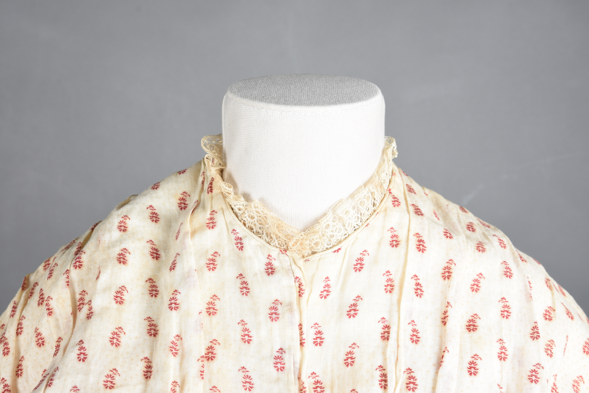 Bomullskjole i to deler, laget av voile med hvit bunn og små strøbuketter i burgunderfarge, hvor prikkene delvis er falmet bort. Skjørtet er sterkt rynket og festet til en smal linning av grov lin, med to legg nederst og en kant med smal bomullsnor. Livet har lange ermer, rund halsringning uten krage, og en skjult hekteåpning som går helt ned. Foran og bak går livet ned i en spiss, med rynkefolder fra skuldrene ned mot spissene. Livet er helfôret, og fôret går inn i ermene. Halsen er kantet med en liten kniplingsrysj, og det er to stoffknapper ved hvert håndledd. Hemper for oppheng er innsydd i skuldersømmene, og det er opphengshemper sydd inn i skjørtets linning. Kjolen er håndsydd.