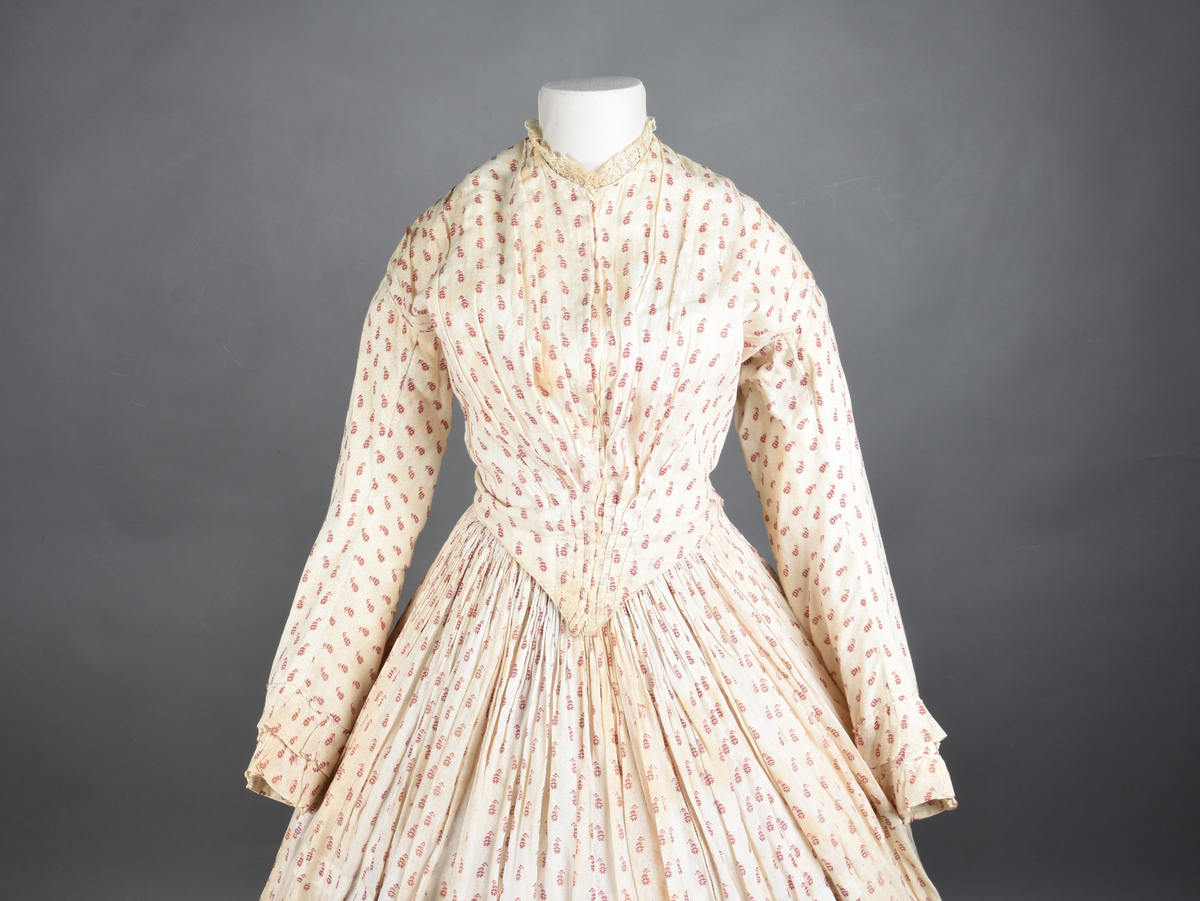 Bomullskjole i to deler, laget av voile med hvit bunn og små strøbuketter i burgunderfarge, hvor prikkene delvis er falmet bort. Skjørtet er sterkt rynket og festet til en smal linning av grov lin, med to legg nederst og en kant med smal bomullsnor. Livet har lange ermer, rund halsringning uten krage, og en skjult hekteåpning som går helt ned. Foran og bak går livet ned i en spiss, med rynkefolder fra skuldrene ned mot spissene. Livet er helfôret, og fôret går inn i ermene. Halsen er kantet med en liten kniplingsrysj, og det er to stoffknapper ved hvert håndledd. Hemper for oppheng er innsydd i skuldersømmene, og det er opphengshemper sydd inn i skjørtets linning. Kjolen er håndsydd.