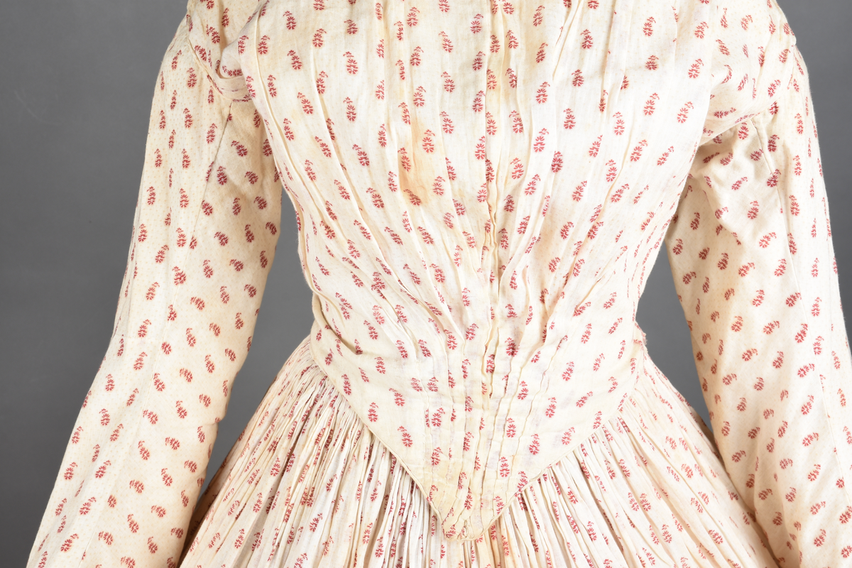 Bomullskjole i to deler, laget av voile med hvit bunn og små strøbuketter i burgunderfarge, hvor prikkene delvis er falmet bort. Skjørtet er sterkt rynket og festet til en smal linning av grov lin, med to legg nederst og en kant med smal bomullsnor. Livet har lange ermer, rund halsringning uten krage, og en skjult hekteåpning som går helt ned. Foran og bak går livet ned i en spiss, med rynkefolder fra skuldrene ned mot spissene. Livet er helfôret, og fôret går inn i ermene. Halsen er kantet med en liten kniplingsrysj, og det er to stoffknapper ved hvert håndledd. Hemper for oppheng er innsydd i skuldersømmene, og det er opphengshemper sydd inn i skjørtets linning. Kjolen er håndsydd.