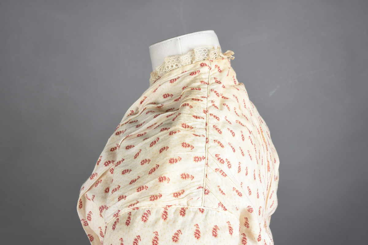 Bomullskjole i to deler, laget av voile med hvit bunn og små strøbuketter i burgunderfarge, hvor prikkene delvis er falmet bort. Skjørtet er sterkt rynket og festet til en smal linning av grov lin, med to legg nederst og en kant med smal bomullsnor. Livet har lange ermer, rund halsringning uten krage, og en skjult hekteåpning som går helt ned. Foran og bak går livet ned i en spiss, med rynkefolder fra skuldrene ned mot spissene. Livet er helfôret, og fôret går inn i ermene. Halsen er kantet med en liten kniplingsrysj, og det er to stoffknapper ved hvert håndledd. Hemper for oppheng er innsydd i skuldersømmene, og det er opphengshemper sydd inn i skjørtets linning. Kjolen er håndsydd.