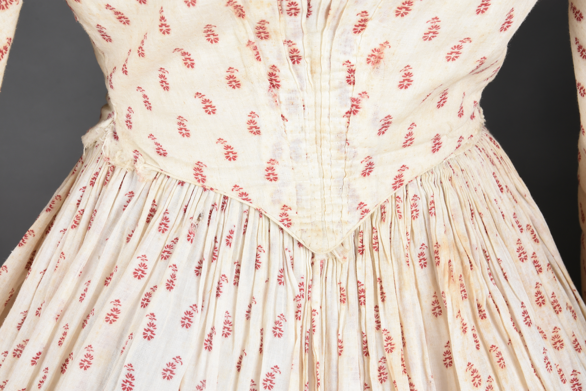 Bomullskjole i to deler, laget av voile med hvit bunn og små strøbuketter i burgunderfarge, hvor prikkene delvis er falmet bort. Skjørtet er sterkt rynket og festet til en smal linning av grov lin, med to legg nederst og en kant med smal bomullsnor. Livet har lange ermer, rund halsringning uten krage, og en skjult hekteåpning som går helt ned. Foran og bak går livet ned i en spiss, med rynkefolder fra skuldrene ned mot spissene. Livet er helfôret, og fôret går inn i ermene. Halsen er kantet med en liten kniplingsrysj, og det er to stoffknapper ved hvert håndledd. Hemper for oppheng er innsydd i skuldersømmene, og det er opphengshemper sydd inn i skjørtets linning. Kjolen er håndsydd.