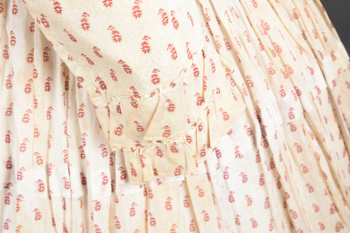 Bomullskjole i to deler, laget av voile med hvit bunn og små strøbuketter i burgunderfarge, hvor prikkene delvis er falmet bort. Skjørtet er sterkt rynket og festet til en smal linning av grov lin, med to legg nederst og en kant med smal bomullsnor. Livet har lange ermer, rund halsringning uten krage, og en skjult hekteåpning som går helt ned. Foran og bak går livet ned i en spiss, med rynkefolder fra skuldrene ned mot spissene. Livet er helfôret, og fôret går inn i ermene. Halsen er kantet med en liten kniplingsrysj, og det er to stoffknapper ved hvert håndledd. Hemper for oppheng er innsydd i skuldersømmene, og det er opphengshemper sydd inn i skjørtets linning. Kjolen er håndsydd.