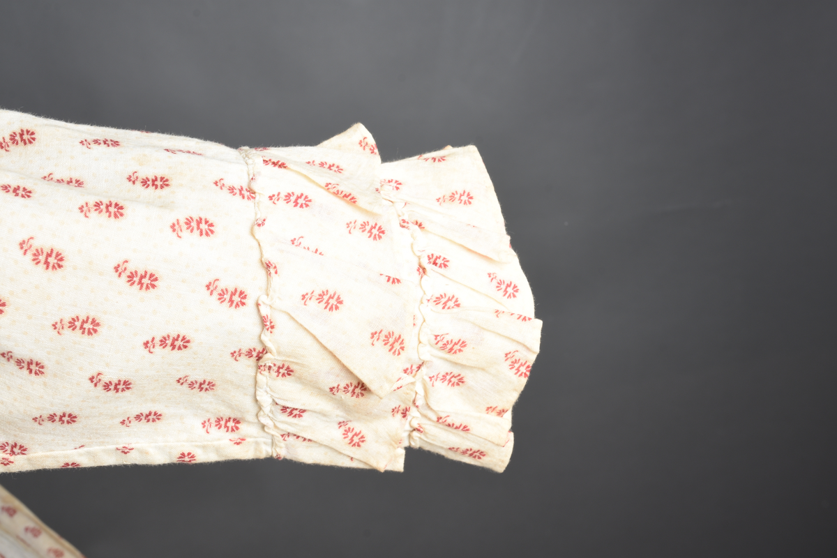 Bomullskjole i to deler, laget av voile med hvit bunn og små strøbuketter i burgunderfarge, hvor prikkene delvis er falmet bort. Skjørtet er sterkt rynket og festet til en smal linning av grov lin, med to legg nederst og en kant med smal bomullsnor. Livet har lange ermer, rund halsringning uten krage, og en skjult hekteåpning som går helt ned. Foran og bak går livet ned i en spiss, med rynkefolder fra skuldrene ned mot spissene. Livet er helfôret, og fôret går inn i ermene. Halsen er kantet med en liten kniplingsrysj, og det er to stoffknapper ved hvert håndledd. Hemper for oppheng er innsydd i skuldersømmene, og det er opphengshemper sydd inn i skjørtets linning. Kjolen er håndsydd.