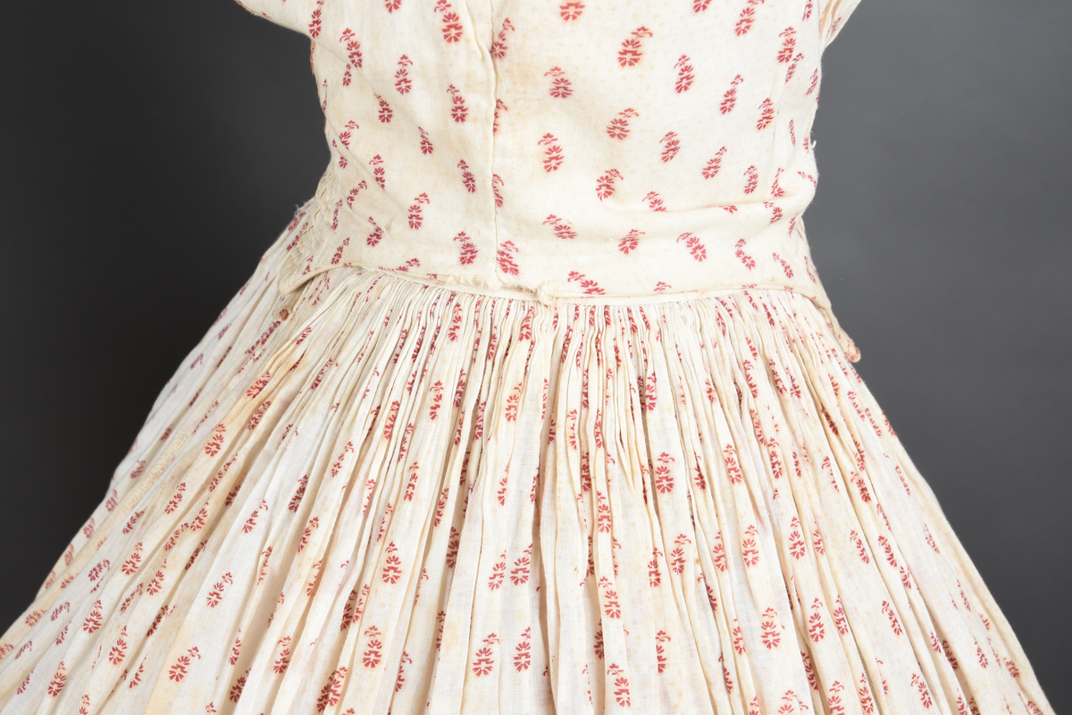 Bomullskjole i to deler, laget av voile med hvit bunn og små strøbuketter i burgunderfarge, hvor prikkene delvis er falmet bort. Skjørtet er sterkt rynket og festet til en smal linning av grov lin, med to legg nederst og en kant med smal bomullsnor. Livet har lange ermer, rund halsringning uten krage, og en skjult hekteåpning som går helt ned. Foran og bak går livet ned i en spiss, med rynkefolder fra skuldrene ned mot spissene. Livet er helfôret, og fôret går inn i ermene. Halsen er kantet med en liten kniplingsrysj, og det er to stoffknapper ved hvert håndledd. Hemper for oppheng er innsydd i skuldersømmene, og det er opphengshemper sydd inn i skjørtets linning. Kjolen er håndsydd.