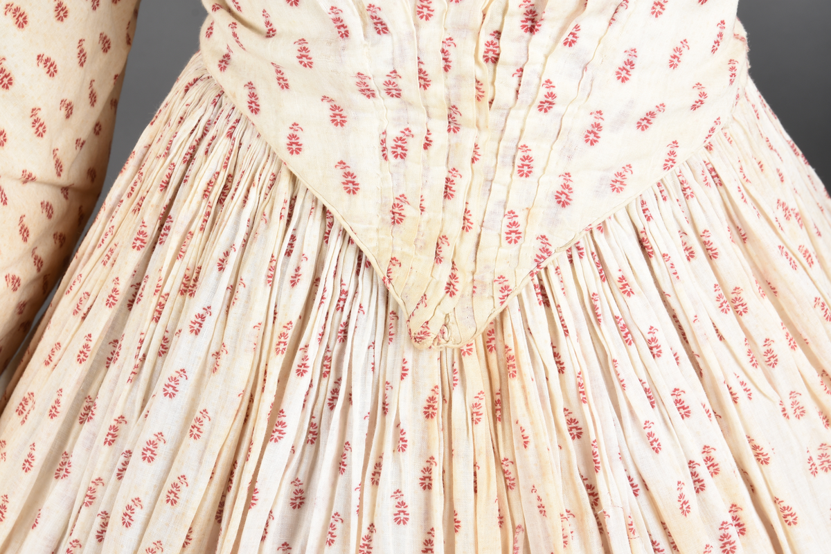 Bomullskjole i to deler, laget av voile med hvit bunn og små strøbuketter i burgunderfarge, hvor prikkene delvis er falmet bort. Skjørtet er sterkt rynket og festet til en smal linning av grov lin, med to legg nederst og en kant med smal bomullsnor. Livet har lange ermer, rund halsringning uten krage, og en skjult hekteåpning som går helt ned. Foran og bak går livet ned i en spiss, med rynkefolder fra skuldrene ned mot spissene. Livet er helfôret, og fôret går inn i ermene. Halsen er kantet med en liten kniplingsrysj, og det er to stoffknapper ved hvert håndledd. Hemper for oppheng er innsydd i skuldersømmene, og det er opphengshemper sydd inn i skjørtets linning. Kjolen er håndsydd.