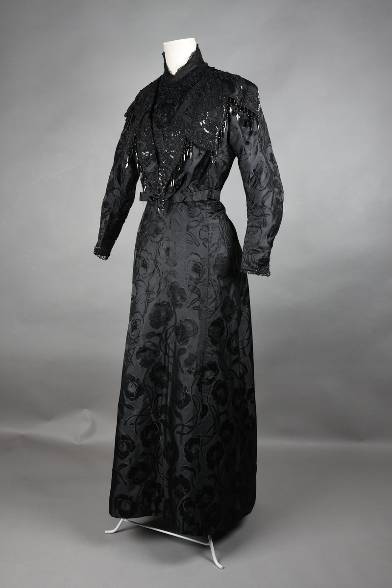 Kjoleliv av sort brokade. Silkeinnfelling og tyllinnfelling foran. Stjernekrave besatt med slag av paljetter og perler. Høy halslinning. Brokaden har et valmuemønster (jugend).