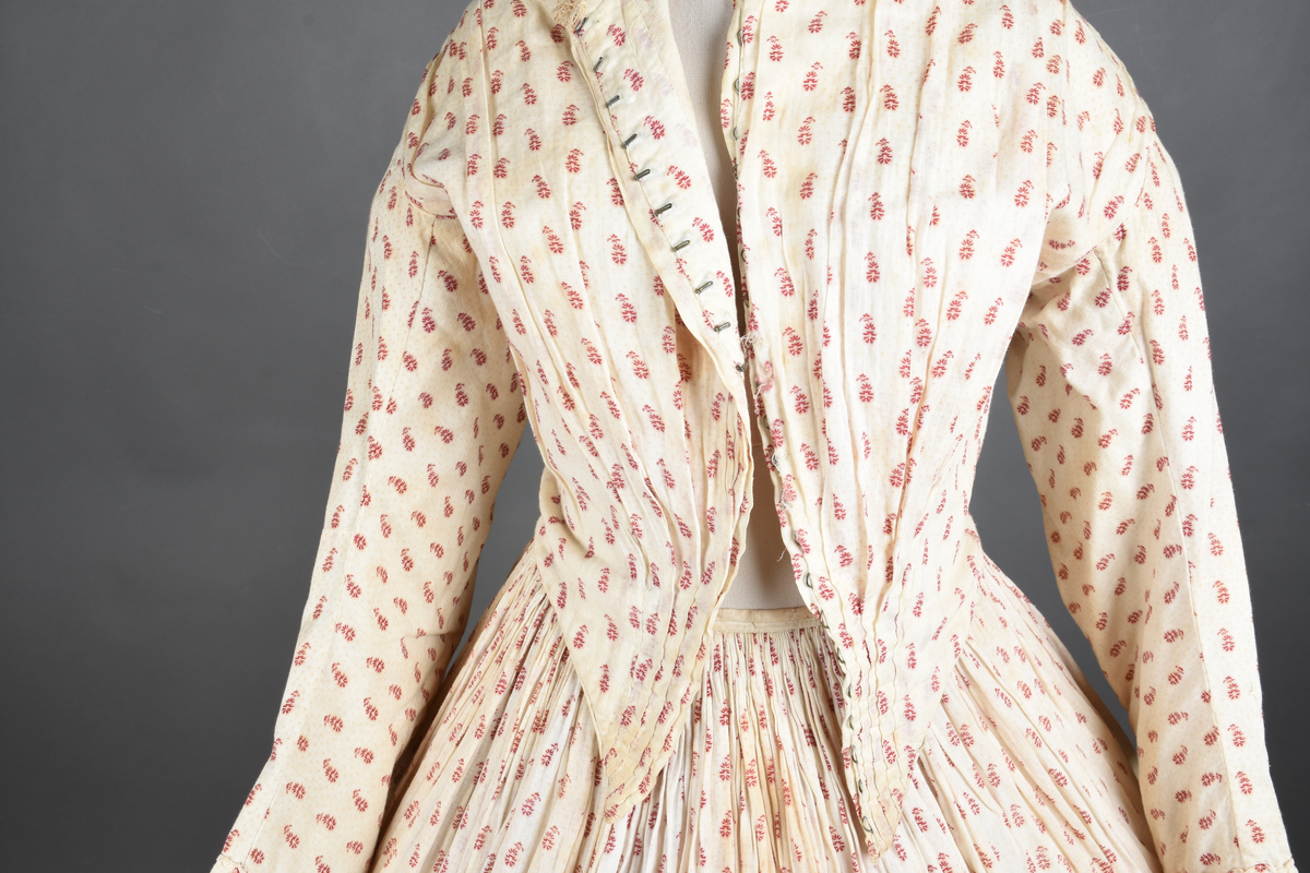 Bomullskjole i to deler, laget av voile med hvit bunn og små strøbuketter i burgunderfarge, hvor prikkene delvis er falmet bort. Skjørtet er sterkt rynket og festet til en smal linning av grov lin, med to legg nederst og en kant med smal bomullsnor. Livet har lange ermer, rund halsringning uten krage, og en skjult hekteåpning som går helt ned. Foran og bak går livet ned i en spiss, med rynkefolder fra skuldrene ned mot spissene. Livet er helfôret, og fôret går inn i ermene. Halsen er kantet med en liten kniplingsrysj, og det er to stoffknapper ved hvert håndledd. Hemper for oppheng er innsydd i skuldersømmene, og det er opphengshemper sydd inn i skjørtets linning. Kjolen er håndsydd.