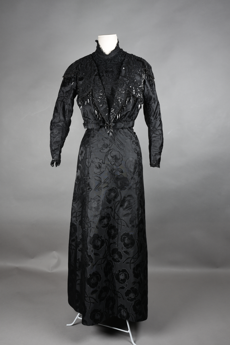 Kjoleliv av sort brokade. Silkeinnfelling og tyllinnfelling foran. Stjernekrave besatt med slag av paljetter og perler. Høy halslinning. Brokaden har et valmuemønster (jugend).