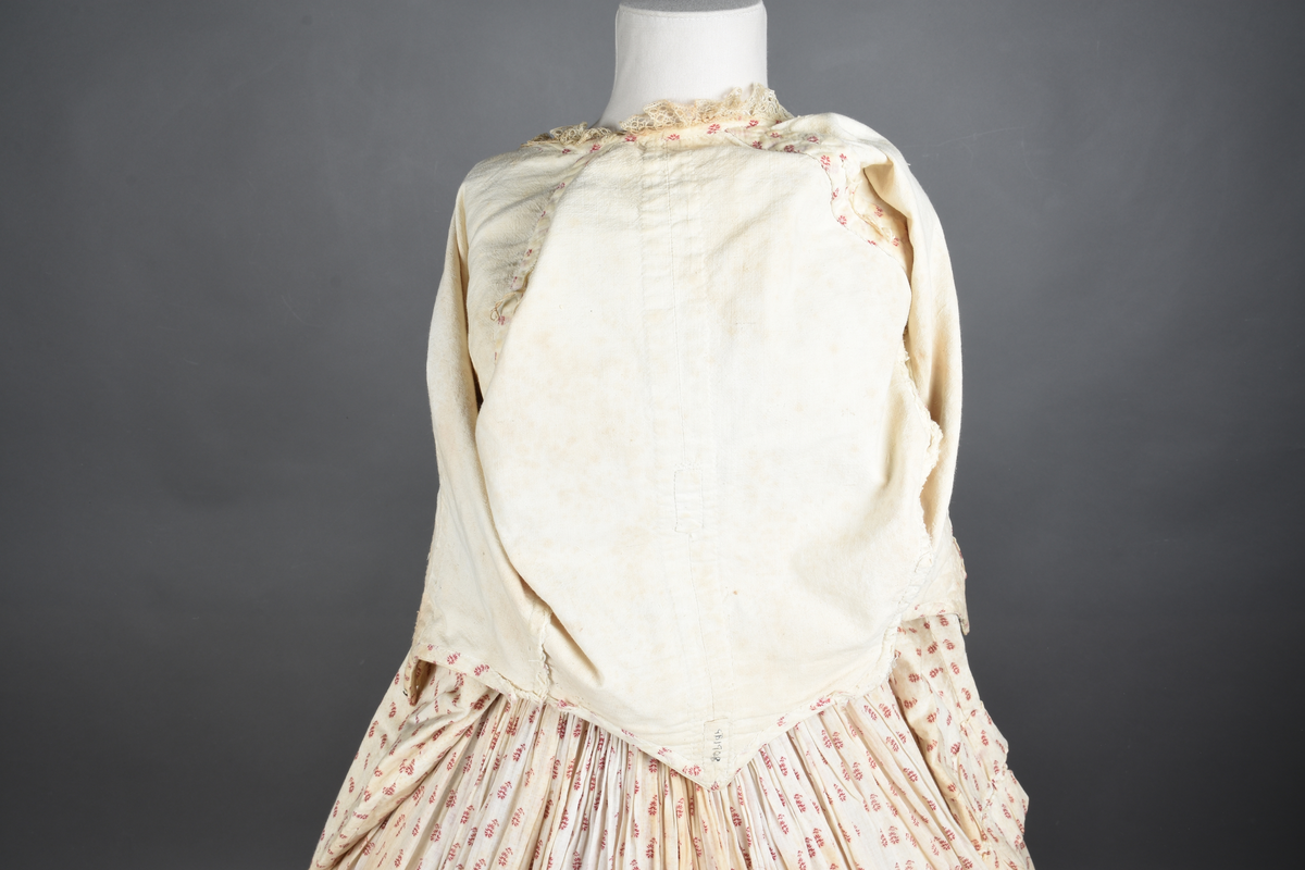 Bomullskjole i to deler, laget av voile med hvit bunn og små strøbuketter i burgunderfarge, hvor prikkene delvis er falmet bort. Skjørtet er sterkt rynket og festet til en smal linning av grov lin, med to legg nederst og en kant med smal bomullsnor. Livet har lange ermer, rund halsringning uten krage, og en skjult hekteåpning som går helt ned. Foran og bak går livet ned i en spiss, med rynkefolder fra skuldrene ned mot spissene. Livet er helfôret, og fôret går inn i ermene. Halsen er kantet med en liten kniplingsrysj, og det er to stoffknapper ved hvert håndledd. Hemper for oppheng er innsydd i skuldersømmene, og det er opphengshemper sydd inn i skjørtets linning. Kjolen er håndsydd.