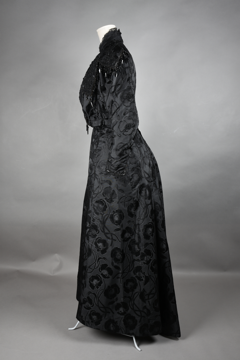 Kjoleliv av sort brokade. Silkeinnfelling og tyllinnfelling foran. Stjernekrave besatt med slag av paljetter og perler. Høy halslinning. Brokaden har et valmuemønster (jugend).