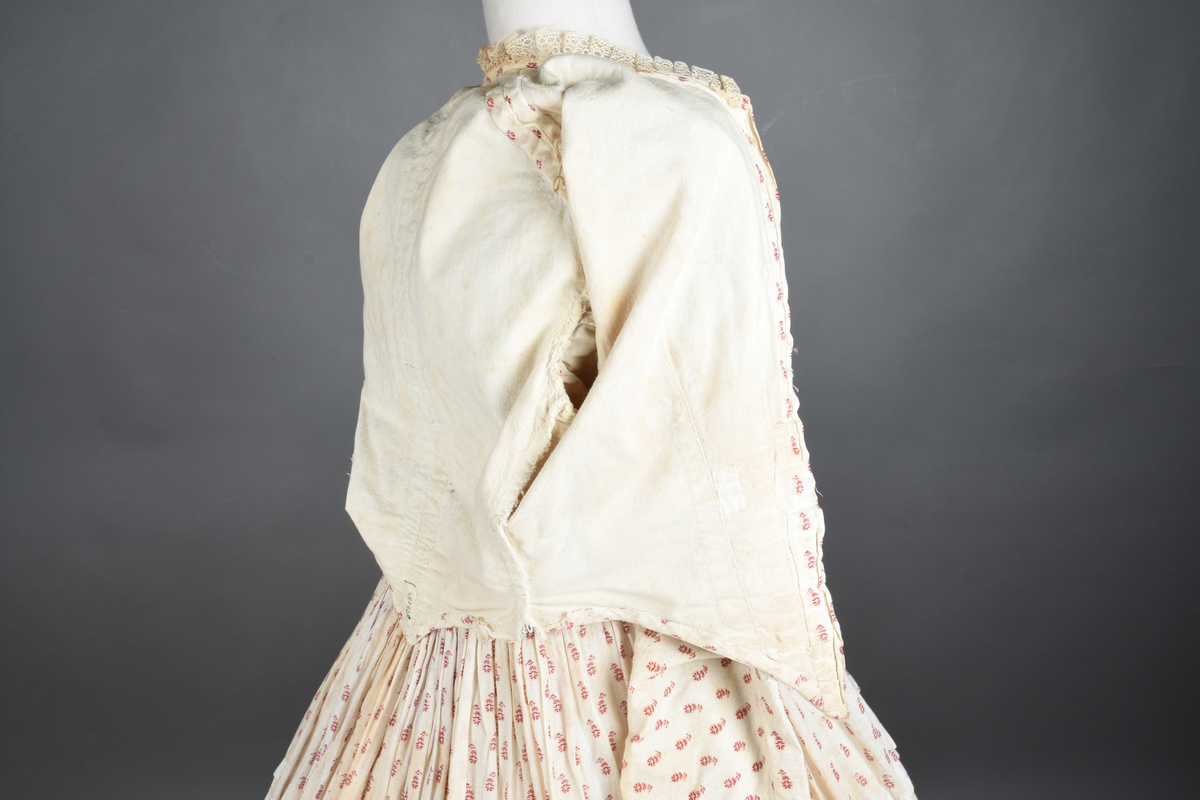 Bomullskjole i to deler, laget av voile med hvit bunn og små strøbuketter i burgunderfarge, hvor prikkene delvis er falmet bort. Skjørtet er sterkt rynket og festet til en smal linning av grov lin, med to legg nederst og en kant med smal bomullsnor. Livet har lange ermer, rund halsringning uten krage, og en skjult hekteåpning som går helt ned. Foran og bak går livet ned i en spiss, med rynkefolder fra skuldrene ned mot spissene. Livet er helfôret, og fôret går inn i ermene. Halsen er kantet med en liten kniplingsrysj, og det er to stoffknapper ved hvert håndledd. Hemper for oppheng er innsydd i skuldersømmene, og det er opphengshemper sydd inn i skjørtets linning. Kjolen er håndsydd.