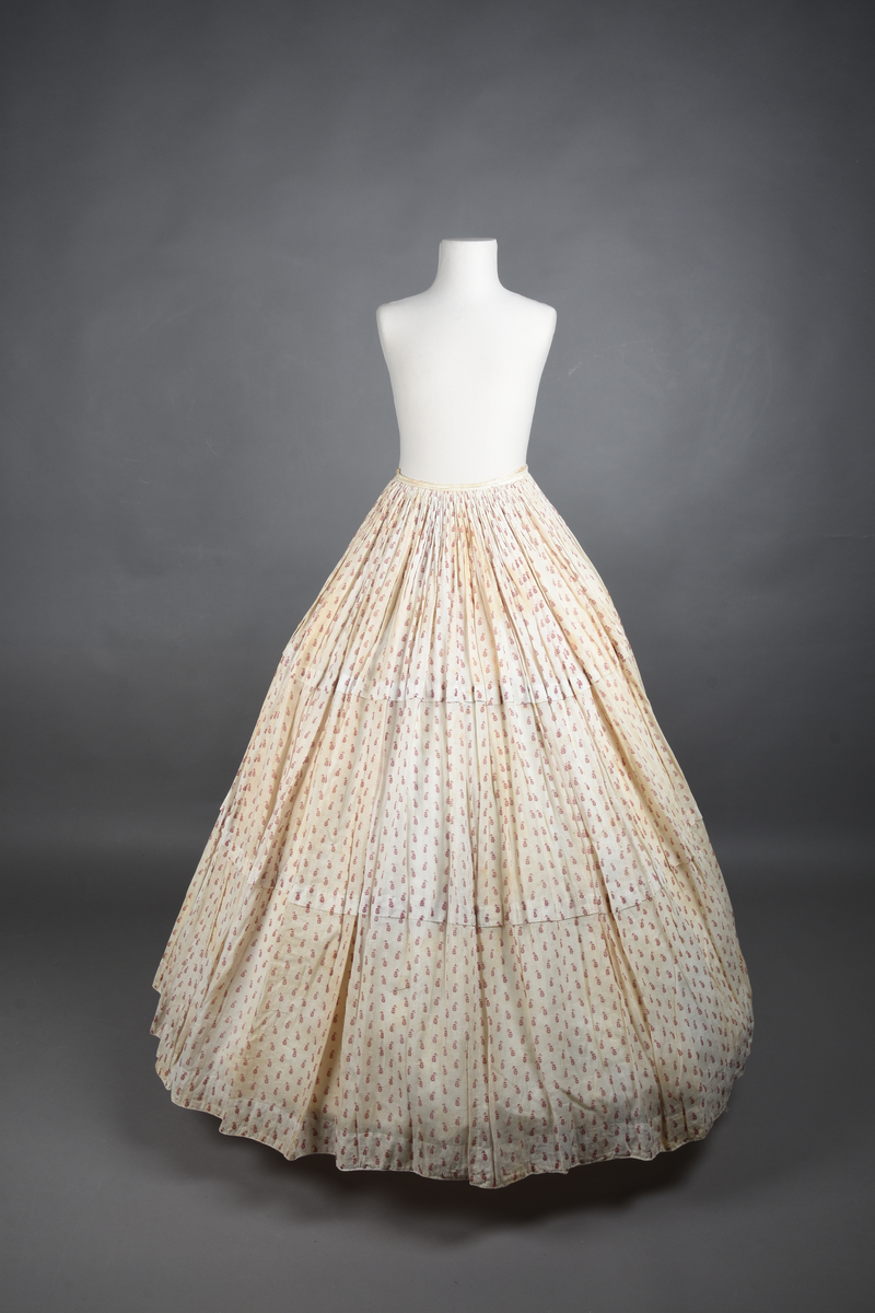 Bomullskjole i to deler, laget av voile med hvit bunn og små strøbuketter i burgunderfarge, hvor prikkene delvis er falmet bort. Skjørtet er sterkt rynket og festet til en smal linning av grov lin, med to legg nederst og en kant med smal bomullsnor. Livet har lange ermer, rund halsringning uten krage, og en skjult hekteåpning som går helt ned. Foran og bak går livet ned i en spiss, med rynkefolder fra skuldrene ned mot spissene. Livet er helfôret, og fôret går inn i ermene. Halsen er kantet med en liten kniplingsrysj, og det er to stoffknapper ved hvert håndledd. Hemper for oppheng er innsydd i skuldersømmene, og det er opphengshemper sydd inn i skjørtets linning. Kjolen er håndsydd.