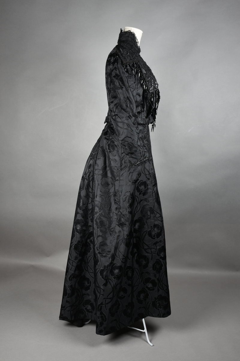 Kjoleliv av sort brokade. Silkeinnfelling og tyllinnfelling foran. Stjernekrave besatt med slag av paljetter og perler. Høy halslinning. Brokaden har et valmuemønster (jugend).