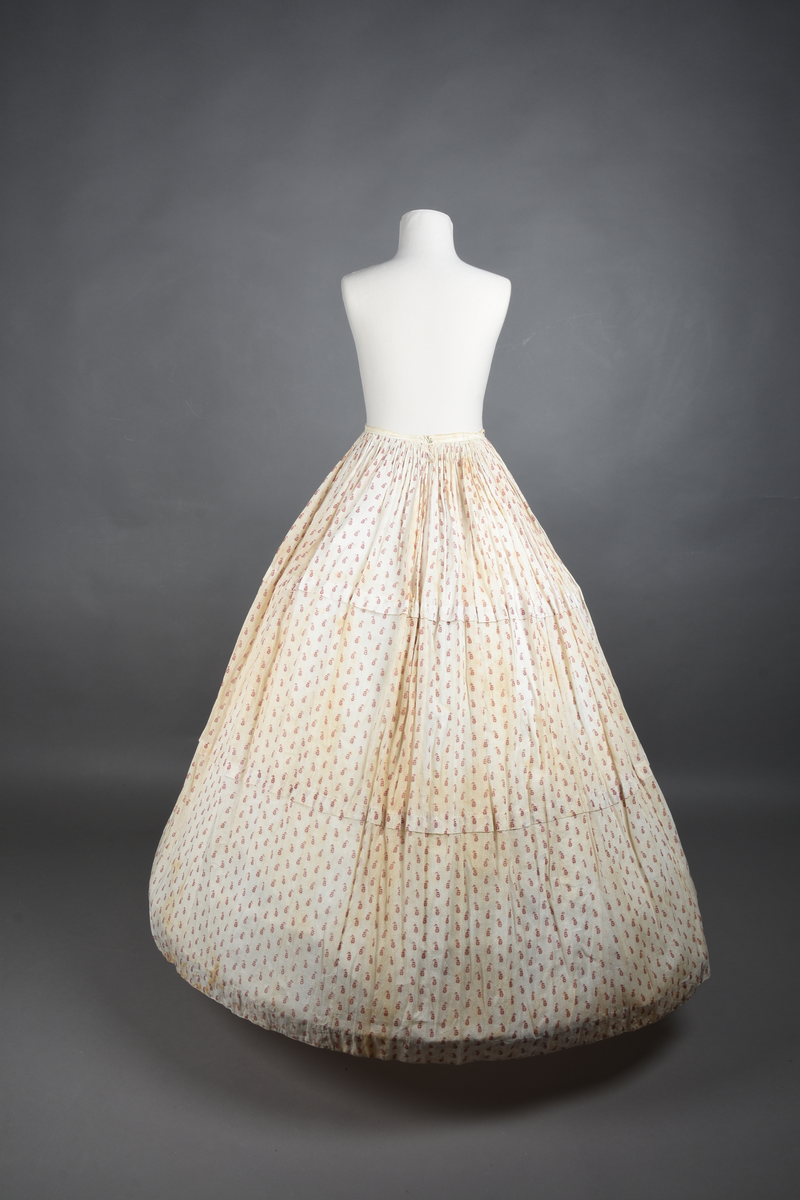 Bomullskjole i to deler, laget av voile med hvit bunn og små strøbuketter i burgunderfarge, hvor prikkene delvis er falmet bort. Skjørtet er sterkt rynket og festet til en smal linning av grov lin, med to legg nederst og en kant med smal bomullsnor. Livet har lange ermer, rund halsringning uten krage, og en skjult hekteåpning som går helt ned. Foran og bak går livet ned i en spiss, med rynkefolder fra skuldrene ned mot spissene. Livet er helfôret, og fôret går inn i ermene. Halsen er kantet med en liten kniplingsrysj, og det er to stoffknapper ved hvert håndledd. Hemper for oppheng er innsydd i skuldersømmene, og det er opphengshemper sydd inn i skjørtets linning. Kjolen er håndsydd.