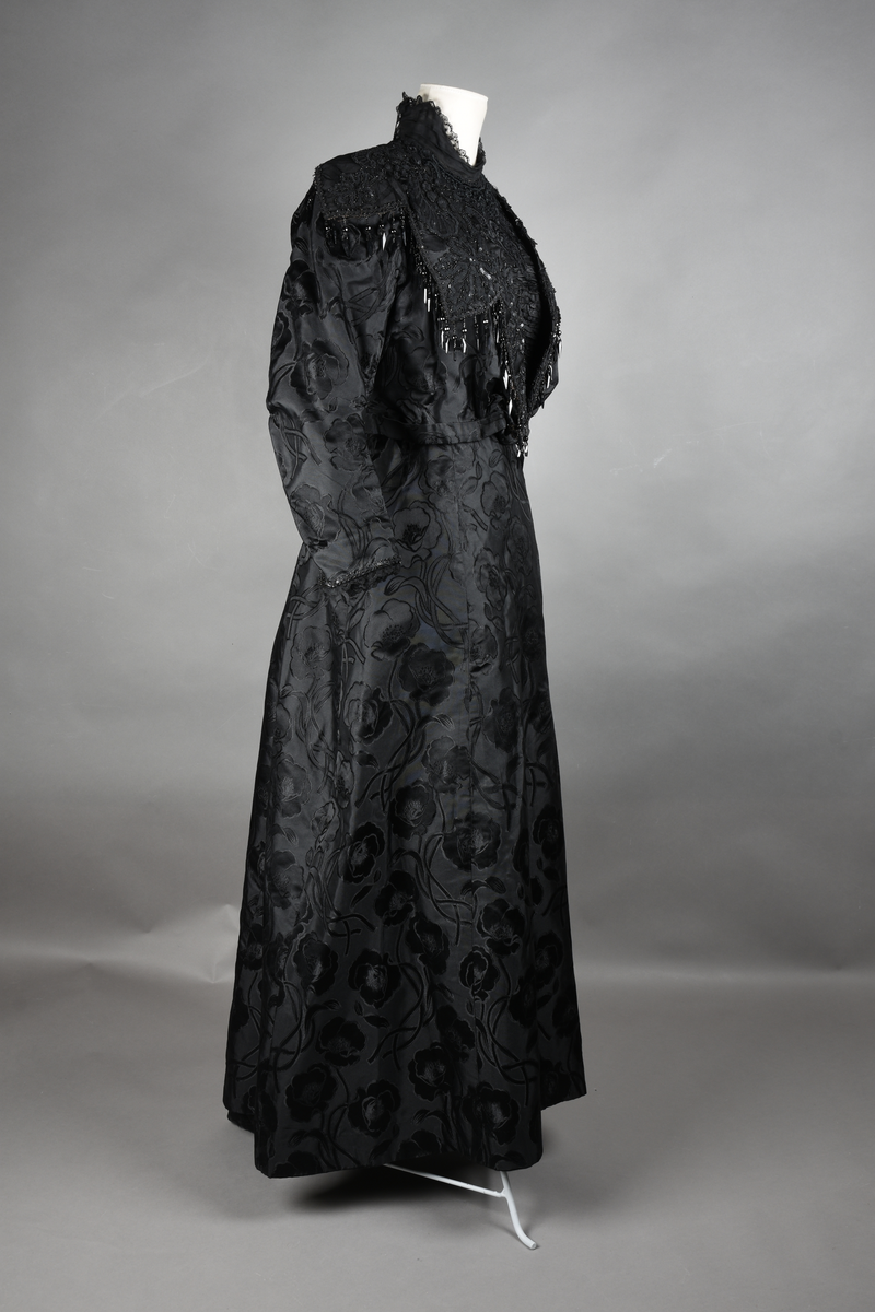 Kjoleliv av sort brokade. Silkeinnfelling og tyllinnfelling foran. Stjernekrave besatt med slag av paljetter og perler. Høy halslinning. Brokaden har et valmuemønster (jugend).