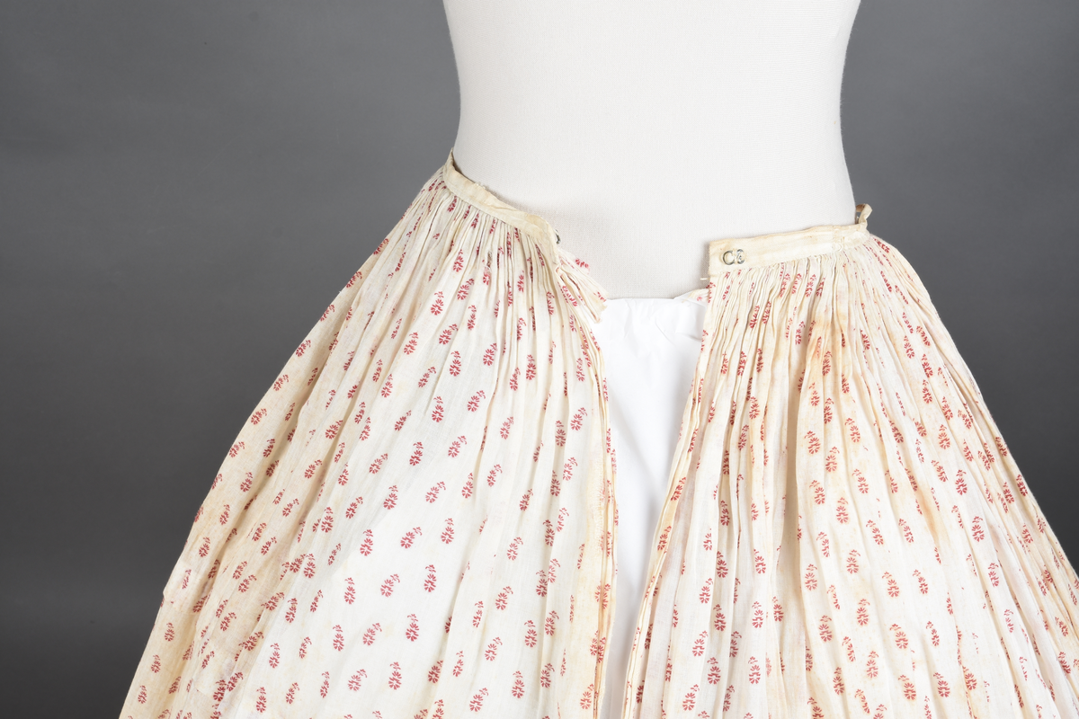 Bomullskjole i to deler, laget av voile med hvit bunn og små strøbuketter i burgunderfarge, hvor prikkene delvis er falmet bort. Skjørtet er sterkt rynket og festet til en smal linning av grov lin, med to legg nederst og en kant med smal bomullsnor. Livet har lange ermer, rund halsringning uten krage, og en skjult hekteåpning som går helt ned. Foran og bak går livet ned i en spiss, med rynkefolder fra skuldrene ned mot spissene. Livet er helfôret, og fôret går inn i ermene. Halsen er kantet med en liten kniplingsrysj, og det er to stoffknapper ved hvert håndledd. Hemper for oppheng er innsydd i skuldersømmene, og det er opphengshemper sydd inn i skjørtets linning. Kjolen er håndsydd.