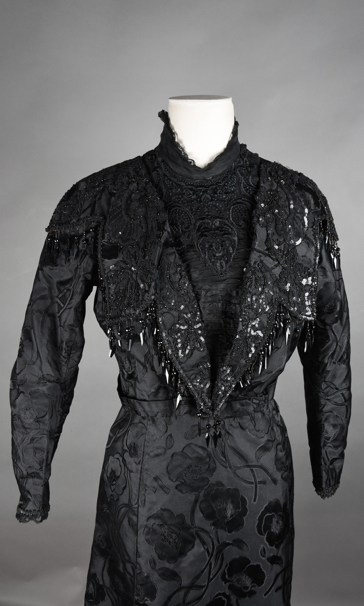 Kjoleliv av sort brokade. Silkeinnfelling og tyllinnfelling foran. Stjernekrave besatt med slag av paljetter og perler. Høy halslinning. Brokaden har et valmuemønster (jugend).