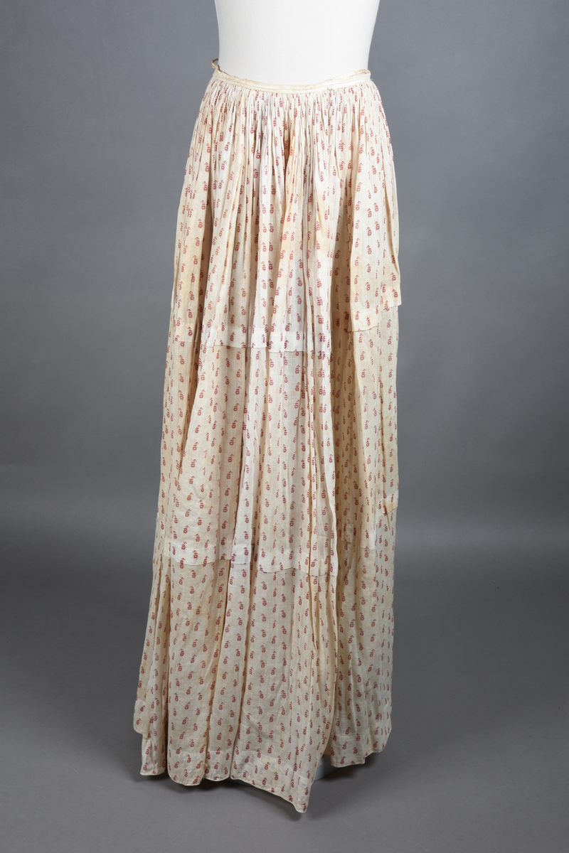 Bomullskjole i to deler, laget av voile med hvit bunn og små strøbuketter i burgunderfarge, hvor prikkene delvis er falmet bort. Skjørtet er sterkt rynket og festet til en smal linning av grov lin, med to legg nederst og en kant med smal bomullsnor. Livet har lange ermer, rund halsringning uten krage, og en skjult hekteåpning som går helt ned. Foran og bak går livet ned i en spiss, med rynkefolder fra skuldrene ned mot spissene. Livet er helfôret, og fôret går inn i ermene. Halsen er kantet med en liten kniplingsrysj, og det er to stoffknapper ved hvert håndledd. Hemper for oppheng er innsydd i skuldersømmene, og det er opphengshemper sydd inn i skjørtets linning. Kjolen er håndsydd.