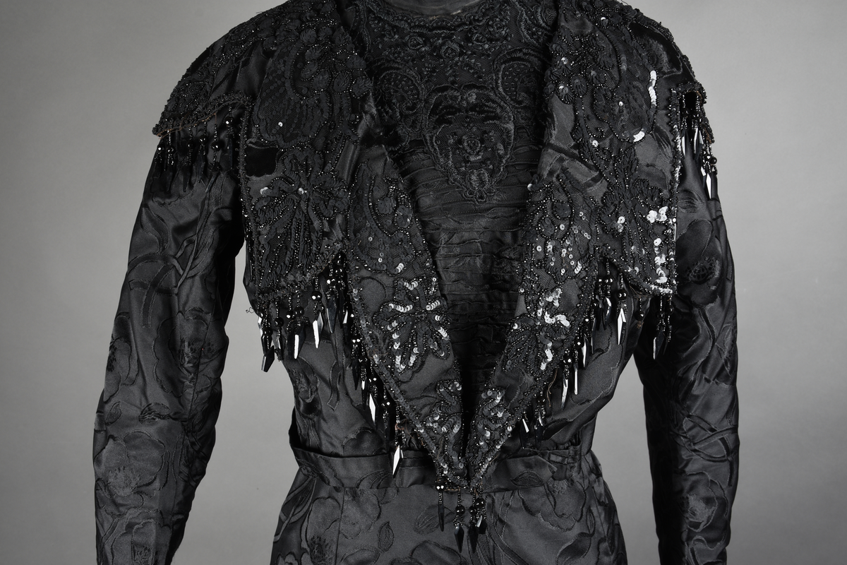 Kjoleliv av sort brokade. Silkeinnfelling og tyllinnfelling foran. Stjernekrave besatt med slag av paljetter og perler. Høy halslinning. Brokaden har et valmuemønster (jugend).