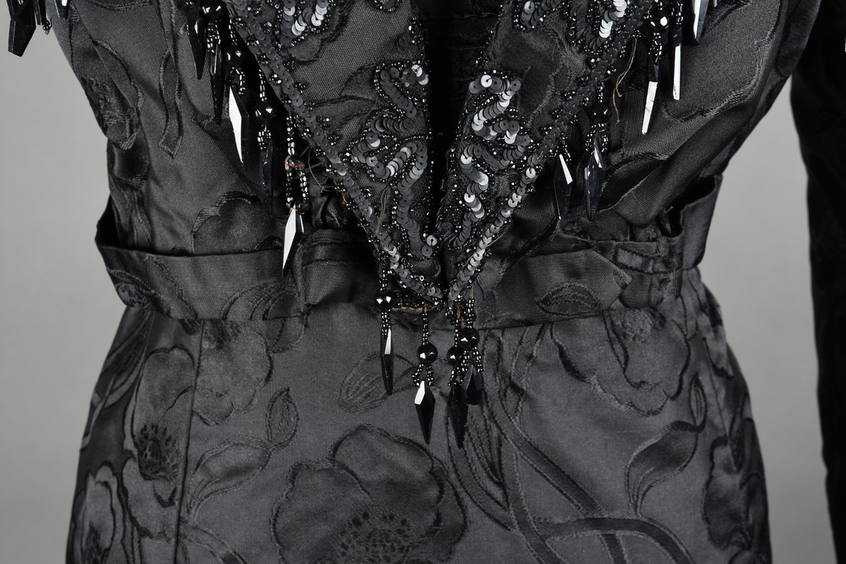 Kjoleliv av sort brokade. Silkeinnfelling og tyllinnfelling foran. Stjernekrave besatt med slag av paljetter og perler. Høy halslinning. Brokaden har et valmuemønster (jugend).