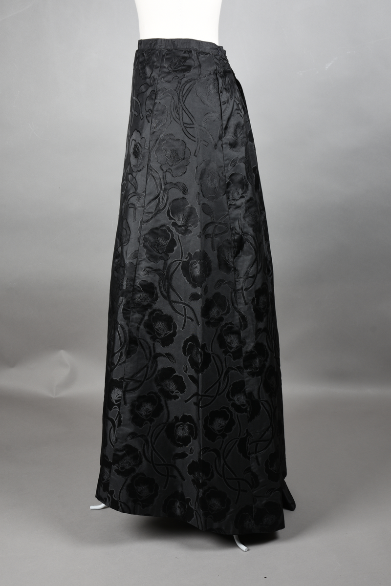 Skjørt av brokade med valmuemønster (jugend). Slep bak og ekstra vidde over setet hvor det er plass til en kø.