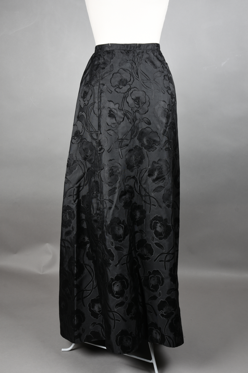 Skjørt av brokade med valmuemønster (jugend). Slep bak og ekstra vidde over setet hvor det er plass til en kø.