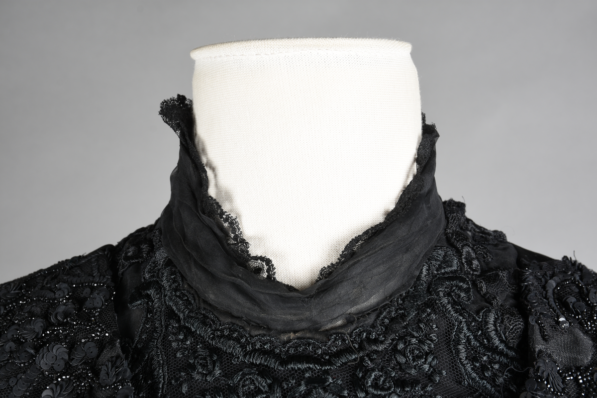 Kjoleliv av sort brokade. Silkeinnfelling og tyllinnfelling foran. Stjernekrave besatt med slag av paljetter og perler. Høy halslinning. Brokaden har et valmuemønster (jugend).