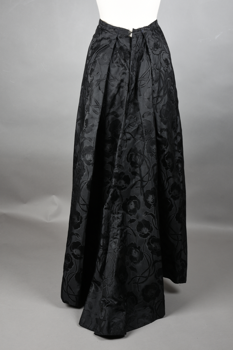 Skjørt av brokade med valmuemønster (jugend). Slep bak og ekstra vidde over setet hvor det er plass til en kø.