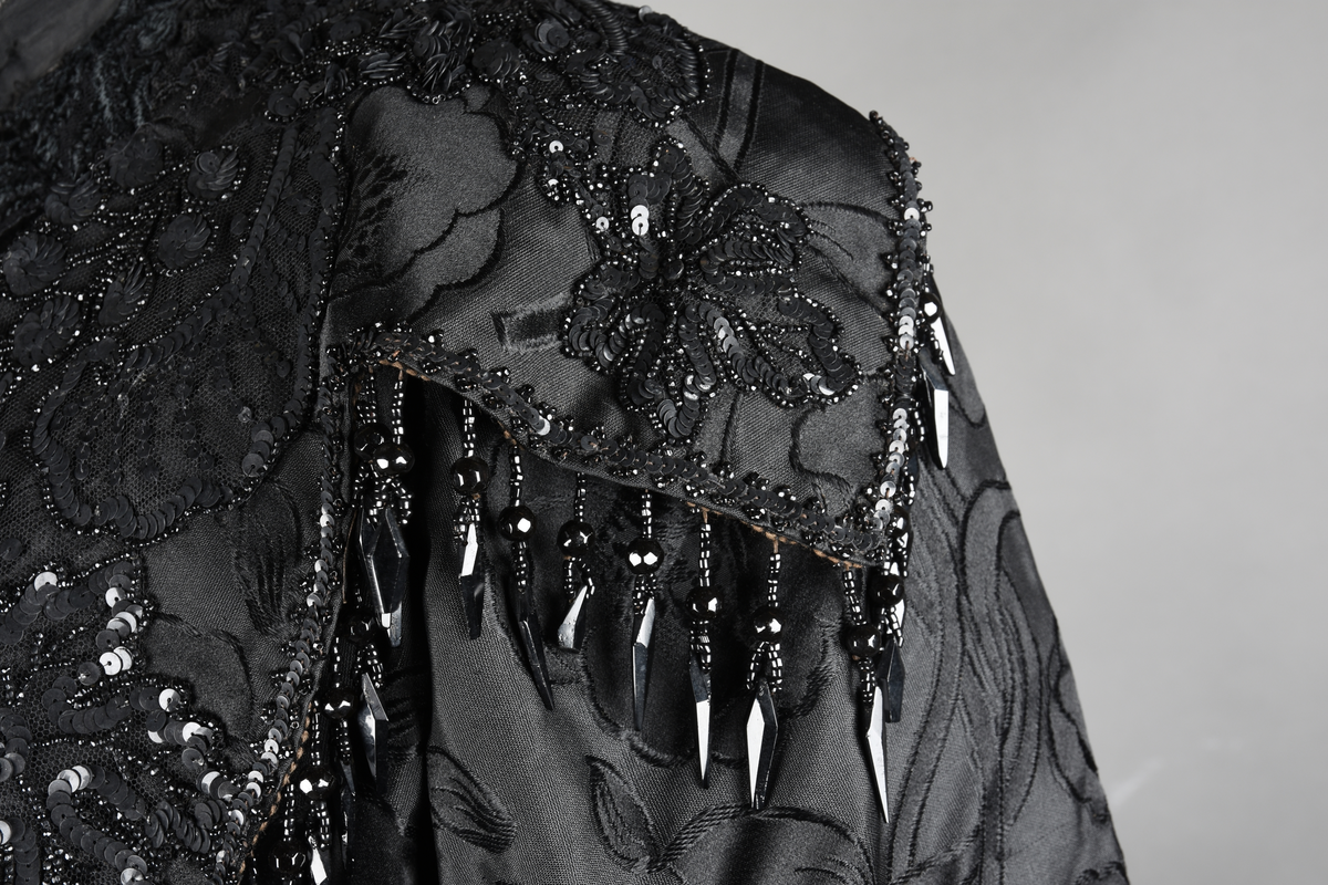 Kjoleliv av sort brokade. Silkeinnfelling og tyllinnfelling foran. Stjernekrave besatt med slag av paljetter og perler. Høy halslinning. Brokaden har et valmuemønster (jugend).