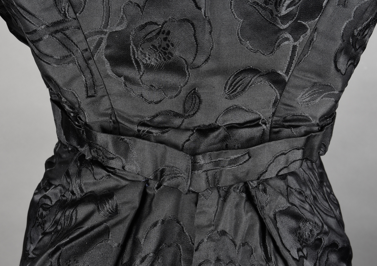 Kjoleliv av sort brokade. Silkeinnfelling og tyllinnfelling foran. Stjernekrave besatt med slag av paljetter og perler. Høy halslinning. Brokaden har et valmuemønster (jugend).