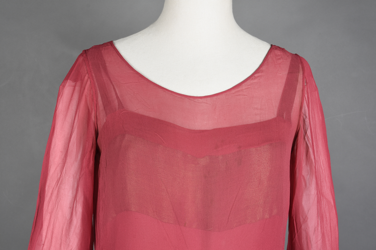 Overkjole av bringebærrød chiffon med garnityr av bronsebrun taft lagt på som skjell og med tre-motiver i tykksømsstrek. I sidene og midt foran oppheftet med bronsebånd (metalltråd?). Langs kanten nede og på ermene er den pyntet med et bredt stykke av brune silkespisser. Halvlange ermer, dyp utringning. Strikk nederst på ermene.