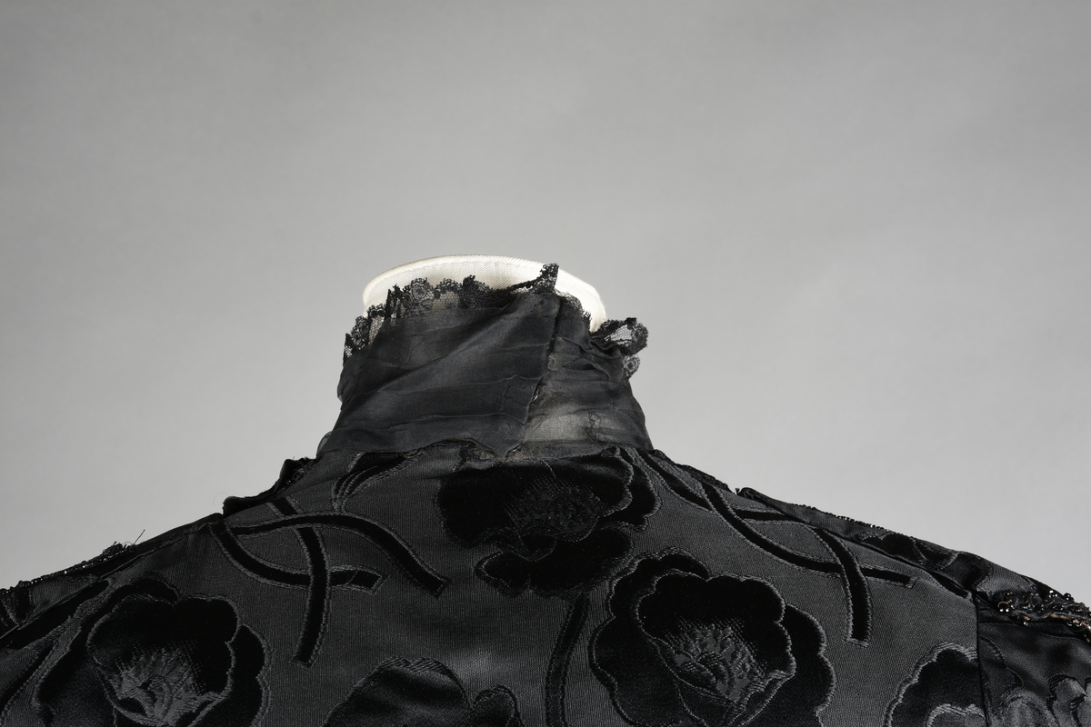 Kjoleliv av sort brokade. Silkeinnfelling og tyllinnfelling foran. Stjernekrave besatt med slag av paljetter og perler. Høy halslinning. Brokaden har et valmuemønster (jugend).