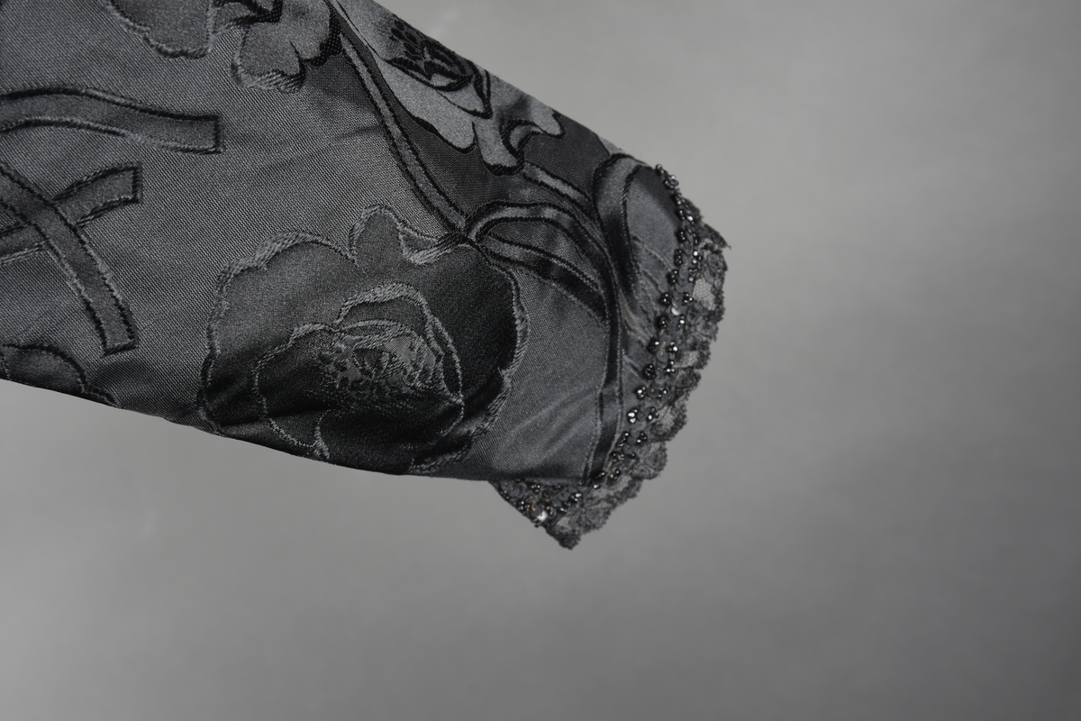 Kjoleliv av sort brokade. Silkeinnfelling og tyllinnfelling foran. Stjernekrave besatt med slag av paljetter og perler. Høy halslinning. Brokaden har et valmuemønster (jugend).