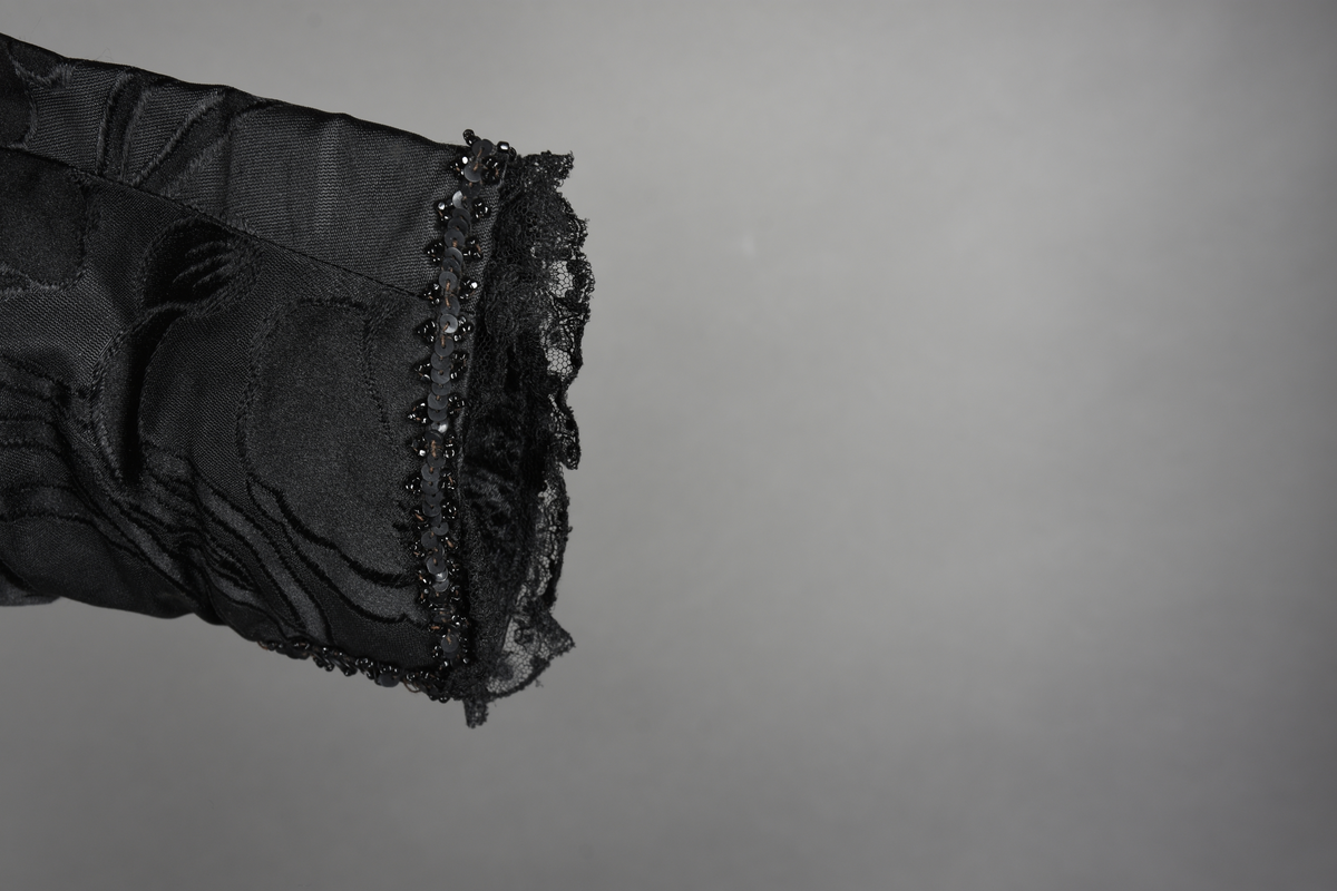 Kjoleliv av sort brokade. Silkeinnfelling og tyllinnfelling foran. Stjernekrave besatt med slag av paljetter og perler. Høy halslinning. Brokaden har et valmuemønster (jugend).