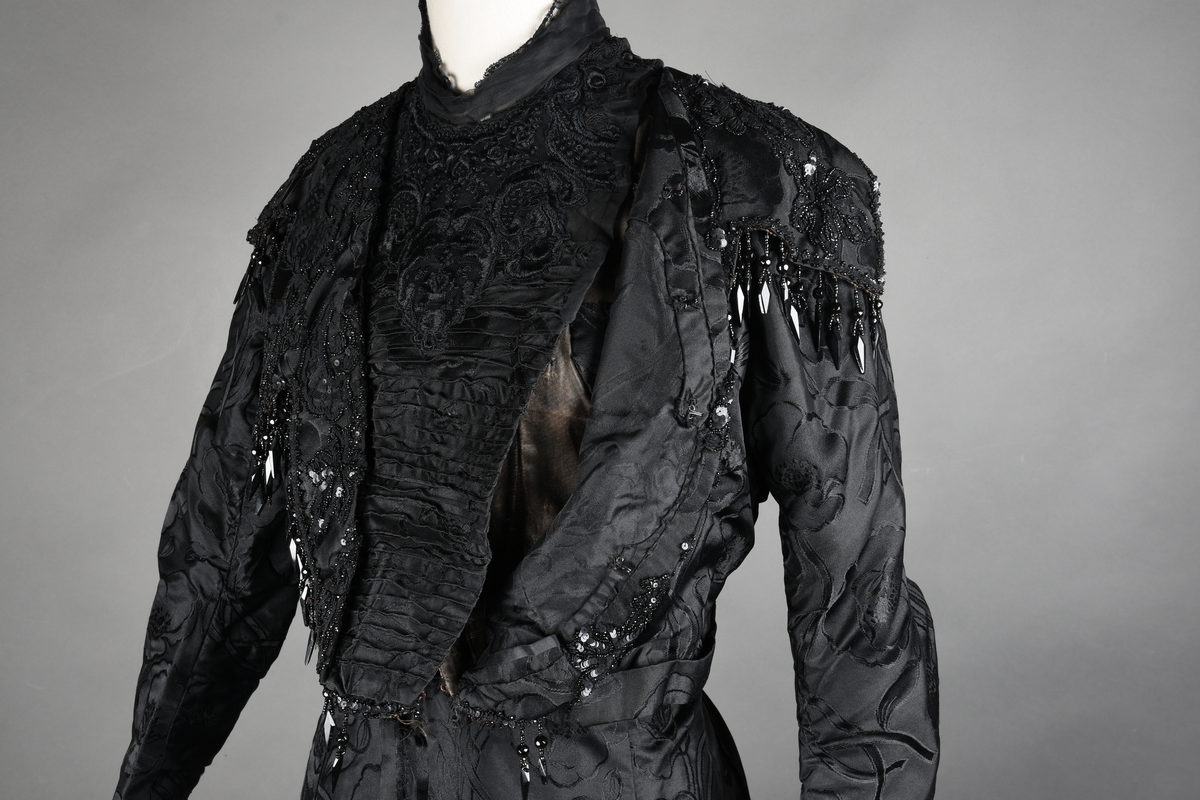 Kjoleliv av sort brokade. Silkeinnfelling og tyllinnfelling foran. Stjernekrave besatt med slag av paljetter og perler. Høy halslinning. Brokaden har et valmuemønster (jugend).